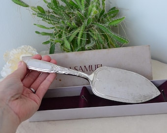 May include: Silver-plated cake server with a decorative handle and a wide, flat blade. The server is presented in a box with the name "SAMUEL" visible. The handle has an oval, white inlay. The server is designed for serving cakes and pastries.