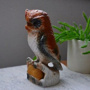 May include: A ceramic owl figurine with brown, white, and black markings. The owl is perched on a base with a yellow cushion.