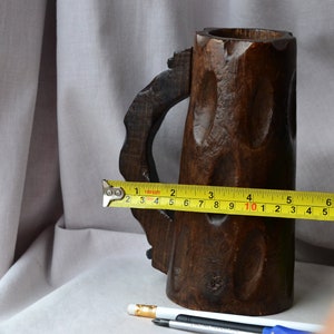 May include: A dark brown wooden mug with a carved handle. The mug is 10 inches tall and is measured with a yellow tape measure.