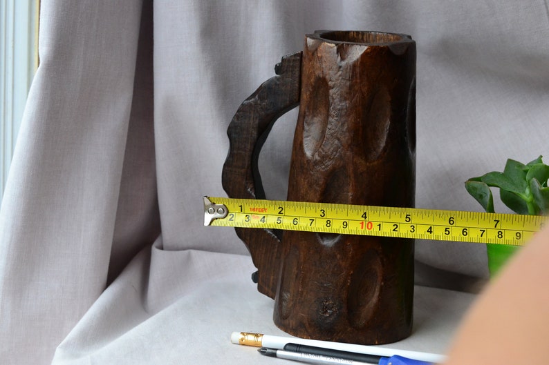 May include: A dark brown wooden mug with a carved handle. The mug is 10 inches tall and is measured with a yellow tape measure.