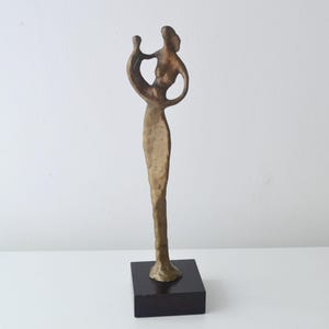 May include: A bronze sculpture depicting two figures in an embrace, set on a black rectangular base. The sculpture has a flowing design and a warm metallic finish. The figures are stylized, with one figure holding the other.