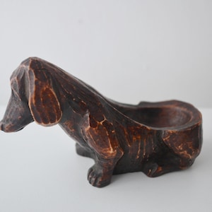 Pode incluir: Estatueta de cão dachshund de madeira entalhada castanha, com uma abertura em forma de tigela nas costas. O cão está sentado, com cabeça e orelhas detalhadas. A madeira tem um aspeto texturizado e rústico.