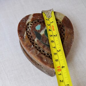 Puede incluir: Una caja de piedra en forma de coraz&oacute;n, de color marr&oacute;n y blanco, con tallas intrincadas e incrustaciones de colores. Una cinta m&eacute;trica amarilla se coloca junto a la caja, con los n&uacute;meros del 1 al 5 visibles.