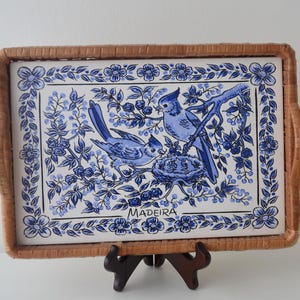 May include: A rectangular serving tray with a woven wicker frame and a central ceramic tile insert. The tile features a blue and white illustration of two birds near a nest, surrounded by floral patterns, with the word MADEIRA printed below.