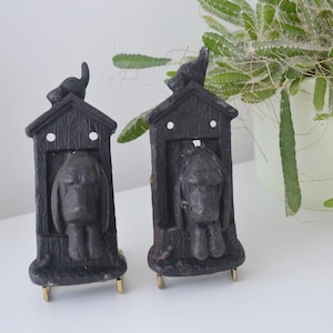 Può includere: Due statue di cani neri in stile vintage. Ogni statua presenta una figura di cane all'interno di una piccola struttura a forma di casa, sormontata da un uccello. Le statue hanno una finitura invecchiata e sono montate su piccoli supporti dorati.