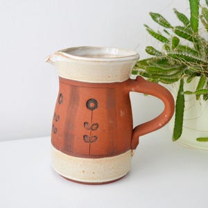 May include: A handmade ceramic jug with a terracotta body and a cream rim and base. The jug features a floral design in black and a curved handle. A decorative piece for the kitchen or dining room.