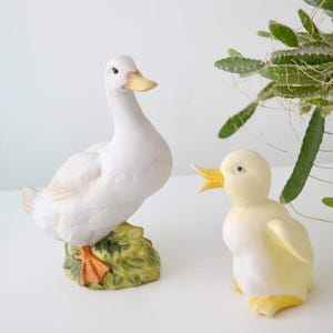 May include: Two ceramic duck figurines. One is a white duck with a yellow beak and orange feet, perched on a green base. The other is a yellow duckling with an open beak. Both are decorative.