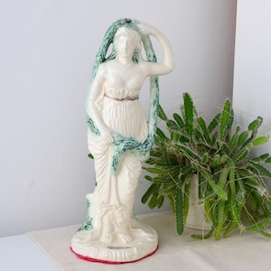 May include: A ceramic statue of a woman in a flowing white gown, with a green and white serpent-like design draped over her head and body. The statue has a red base and a gold-colored belt. A potted plant is in the background.
