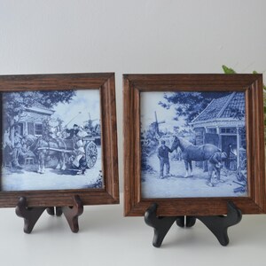 Può includere: Due piastrelle in ceramica blu e bianca incorniciate raffiguranti scene con cavalli e persone. Le piastrelle sono in cornici di legno marrone ed esposte su supporti neri. L'opera d'arte ha un'estetica vintage e rustica.
