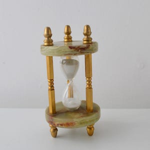 Op de afbeelding: Een zandloper met een doorzichtige glazen bol gevuld met wit zand, ondersteund door een frame met twee ronde groene onyx stenen platen en vier goudkleurige metalen zuilen. De boven- en onderkant van het frame zijn afgewerkt met gouden knoppen.