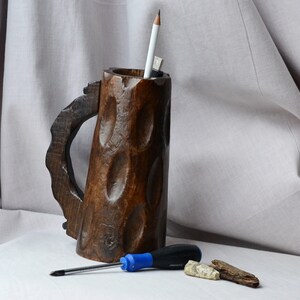 May include: A wooden mug with a carved handle and a rough, textured surface. The mug is used as a pencil holder and contains a pencil and a pen. A blue screwdriver and two pieces of wood are on a white surface in front of the mug.