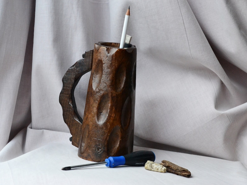 May include: A wooden mug with a carved handle and a rough, textured surface. The mug is used as a pencil holder and contains a pencil and a pen. A blue screwdriver and two pieces of wood are on a white surface in front of the mug.