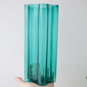 Può includere: Un vaso alto in vetro color teal con un design ondulato unico. Il vaso ha una forma rettangolare con scanalature verticali, creando un aspetto strutturato. Il colore teal è vibrante, rendendolo un pezzo decorativo.