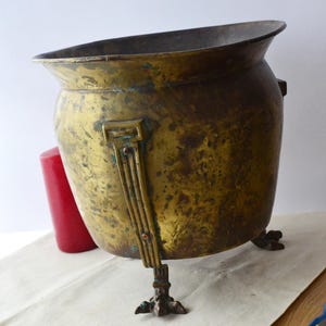 May include: A large, antique brass planter with a rounded body and three decorative feet. The planter has a worn, aged patina and is in good condition.
