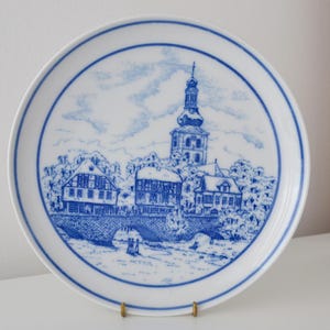 May include: A white ceramic plate with a blue and white illustration of a European town scene. The plate features a detailed depiction of buildings, a bridge, and a church tower, all within a blue border.