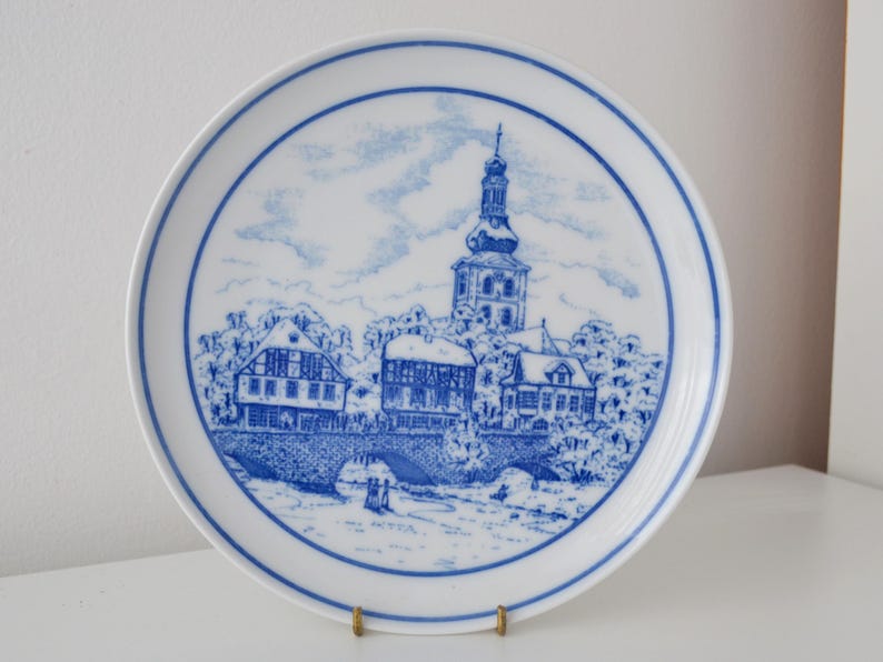 May include: A white ceramic plate with a blue and white illustration of a European town scene. The plate features a detailed depiction of buildings, a bridge, and a church tower, all within a blue border.