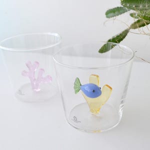 May include: Two clear glass tumblers with decorative sea life inside. One tumbler has a pink coral design, and the other has a blue fish with yellow coral. The glasses are on a white surface.
