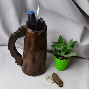 May include: A wooden mug with a handle, carved with a rough texture, used as a desk organizer. It holds a pen, pencil, and a USB drive. A small green pot with a succulent plant sits next to the mug.