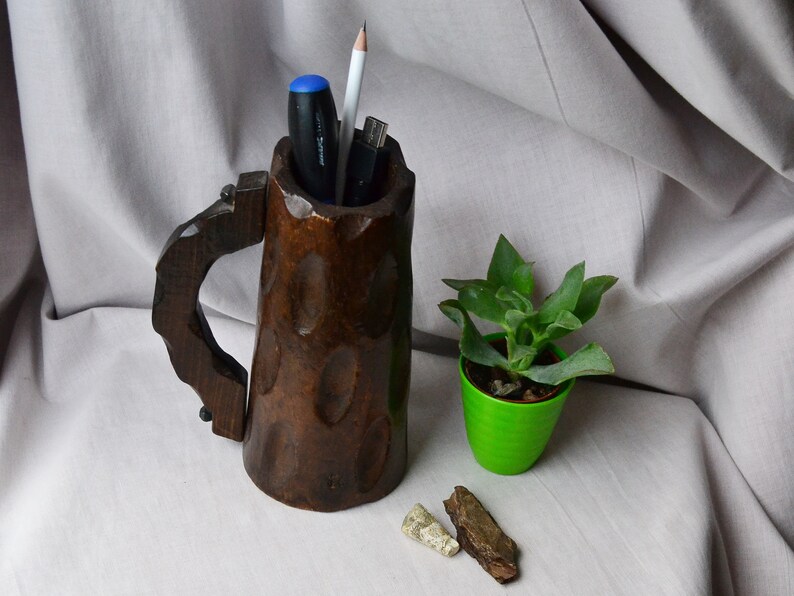 May include: A wooden mug with a handle, carved with a rough texture, used as a desk organizer. It holds a pen, pencil, and a USB drive. A small green pot with a succulent plant sits next to the mug.
