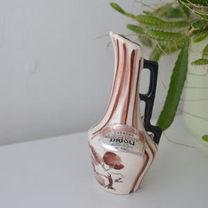 Puede incluir: Pequeña botella de cerámica vintage con base crema y rayas verticales marrones. Tiene un asa negra y una etiqueta que dice "Curaçao Drioli". Ilustraciones florales adornan el frente. La botella es probablemente un artículo decorativo o una botella de licor en miniatura.