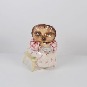 May include: A ceramic hedgehog figurine wearing a red and white patterned dress and apron, sitting on a small bench. The hedgehog holds a small, silver-coloured object. The figurine has a brown head and is painted with fine details.