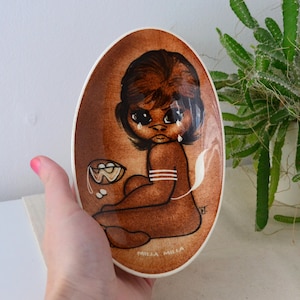 Puede incluir: Plato ovalado de cerámica con diseño marrón y blanco. La obra de arte representa una figura sentada con expresión triste, lágrimas y el texto "Milla Milla". El plato tiene un borde blanco y un fondo marrón.