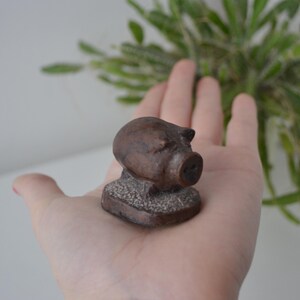 Può includere: Piccola statuetta di maiale marrone con muso e orecchie dettagliate. Il maiale è montato su una base rettangolare strutturata. La statuetta è un oggetto decorativo, probabilmente per una casa o un ufficio.