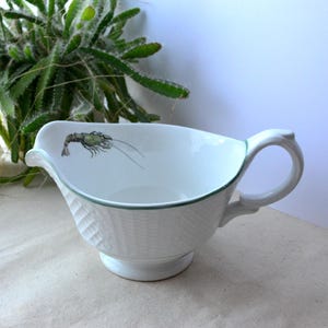 Puede incluir: Salsera de cerámica blanca con borde y asa verdes. El recipiente presenta un patrón texturizado en el exterior y una ilustración pintada de una langosta verde. Un plato de servicio clásico para salsas y jugos.