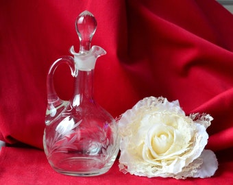 May include: A clear glass bottle with a stopper and etched floral design, sitting on a red surface next to a white fabric rose.