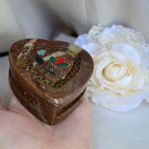Puede incluir: Una caja de piedra de coraz&oacute;n marr&oacute;n con un dise&ntilde;o floral incrustado con piedras de colores. La caja tiene un patr&oacute;n de celos&iacute;a en la parte delantera y est&aacute; abierta.