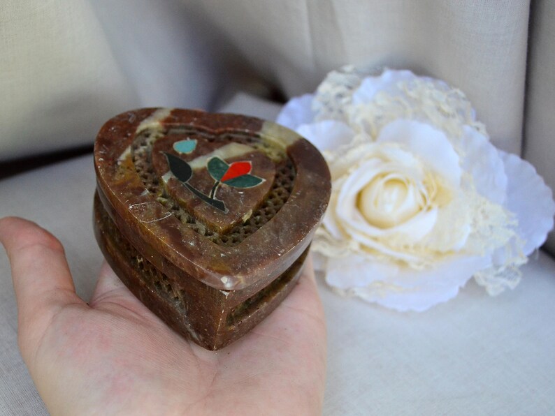 Puede incluir: Una caja de piedra de coraz&oacute;n marr&oacute;n con un dise&ntilde;o floral incrustado con piedras de colores. La caja tiene un patr&oacute;n de celos&iacute;a en la parte delantera y est&aacute; abierta.