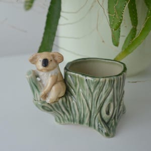 Puede incluir: Una maceta de cerámica con forma de koala. La maceta tiene forma de tocón de árbol de color verde claro. Una pequeña figura de koala está posada en el lateral del tocón. El koala es de color canela con nariz y orejas negras.
