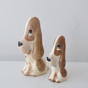 May include: Two ceramic dog figurines in a sitting position, featuring a glossy finish with cream and brown patches. Each dog has long, drooping ears, a black nose, and large blue eyes. The figurines are of different sizes, with one larger than the other.
