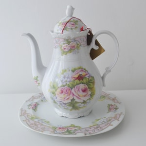 Peut inclure: Une théière en porcelaine blanche avec une assiette ronde assortie, ornée d'un motif floral de roses roses et de feuilles vertes. La théière possède une anse incurvée, un bec verseur et un couvercle avec un petit bouton. Une étiquette brune est attachée.