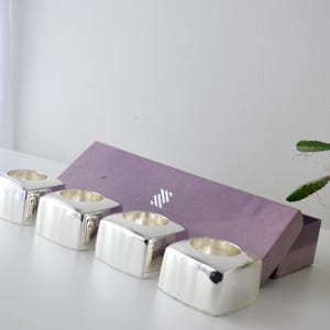 May include: Four silver square candle holders with a circular recess for candles. A purple box with a white logo is behind the candle holders. The candle holders are on a white surface.