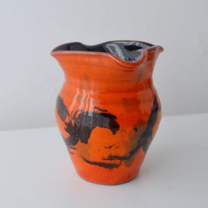 May include: A ceramic pitcher with a glossy orange glaze and dark brown abstract brushstroke patterns. The vessel features a wide, undulating rim and a deep, dark interior, showcasing a rounded body that tapers toward a flat, circular base.