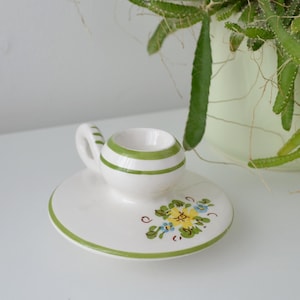 May include: A white ceramic candle holder with a small handle and a round base. The holder is decorated with green stripes and floral designs. A green rim and floral accents add a touch of color. A potted plant is in the background.