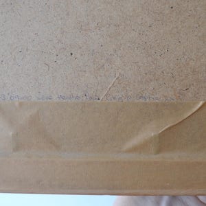 May include: The back of a framed object featuring a light brown paper backing secured to a wooden frame. A rectangular section of the inner fiberboard is visible. Handwritten text in blue ink is present along the lower edge of the fiberboard.
