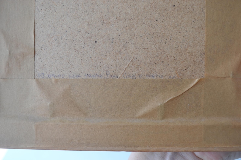 May include: The back of a framed object featuring a light brown paper backing secured to a wooden frame. A rectangular section of the inner fiberboard is visible. Handwritten text in blue ink is present along the lower edge of the fiberboard.