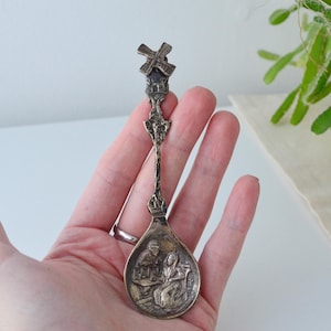 May include: A vintage silver-toned spoon with a windmill design at the top and a detailed scene in the bowl. The spoon is held in a hand, showcasing its intricate craftsmanship and historical charm. A collectible souvenir.