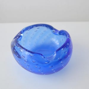 May include: A round, cobalt blue glass ashtray with a bubbly texture. The ashtray has a smooth, curved rim and a deep bowl. The glass is translucent, allowing light to pass through. This vintage ashtray is a decorative item.