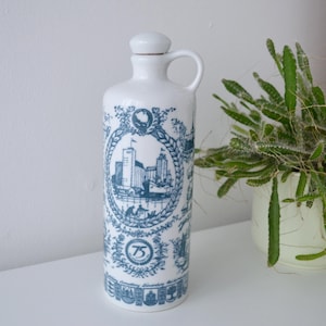 Könnte beinhalten: Weiße Keramikflasche mit Henkel und Verschluss, verziert mit blauen Illustrationen einer Stadtlandschaft und der Zahl 75. Die Flasche ist zylindrisch und steht aufrecht. Eine grüne Pflanze in einem weißen Topf befindet sich im Hintergrund.