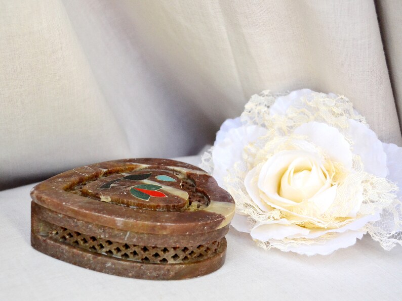 Puede incluir: Una caja de piedra de coraz&oacute;n marr&oacute;n con una incrustaci&oacute;n decorativa de piedras de colores. La caja tiene un dise&ntilde;o de celos&iacute;a alrededor del borde.