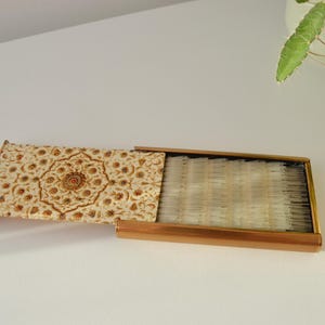 May include: An open, rectangular matchbox with a decorative lid. The lid features a cream-colored background with a detailed floral and geometric pattern in shades of brown and gold. The matchbox is made of metal.