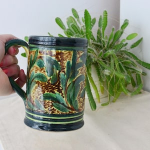 May include: A ceramic mug with a dark green handle and rim, featuring a botanical design in shades of green, brown, and yellow. The mug is held by a hand, showcasing the detailed floral pattern. A plant is in the background.