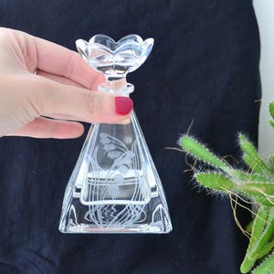 Peut inclure: Un flacon de parfum en verre transparent avec un motif floral. Le flacon a une forme triangulaire et un bouchon en forme de fleur. Le flacon est tenu dans une main sur un fond sombre.