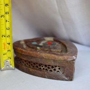 Puede incluir: Una caja de piedra de color marr&oacute;n en forma de coraz&oacute;n con un dise&ntilde;o tallado y piedras de colores incrustadas. La caja tiene un patr&oacute;n de celos&iacute;a en la parte inferior.
