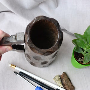May include: A wooden beer stein with a hinged lid and a handle. The stein is dark brown and has a textured surface. The stein is empty and is sitting on a white surface.