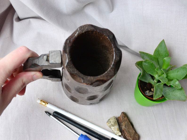 May include: A wooden beer stein with a hinged lid and a handle. The stein is dark brown and has a textured surface. The stein is empty and is sitting on a white surface.
