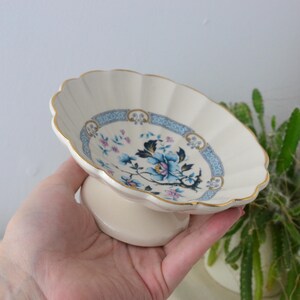 May include: A cream-coloured, scalloped-edge dish with a floral design. The dish has a blue and gold border and a central floral pattern in blue, pink, and white. It is supported by a short, rounded base, likely made of porcelain.